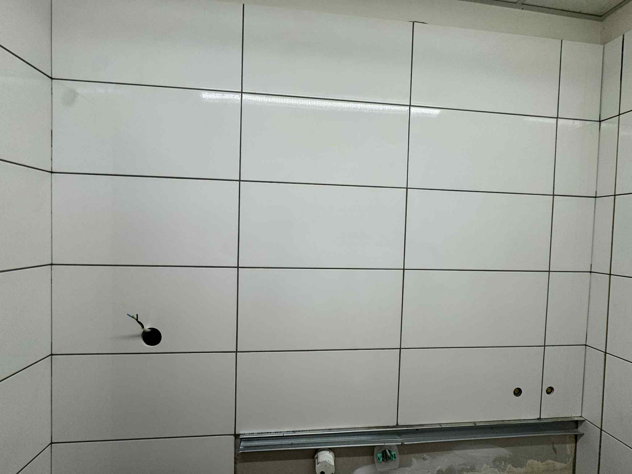 Ściana wyłożona białymi, prostokątnymi płytkami z ciemną fugą. Widoczne otwory na instalacje i listwa przypodłogowa. Minimalistyczny design.