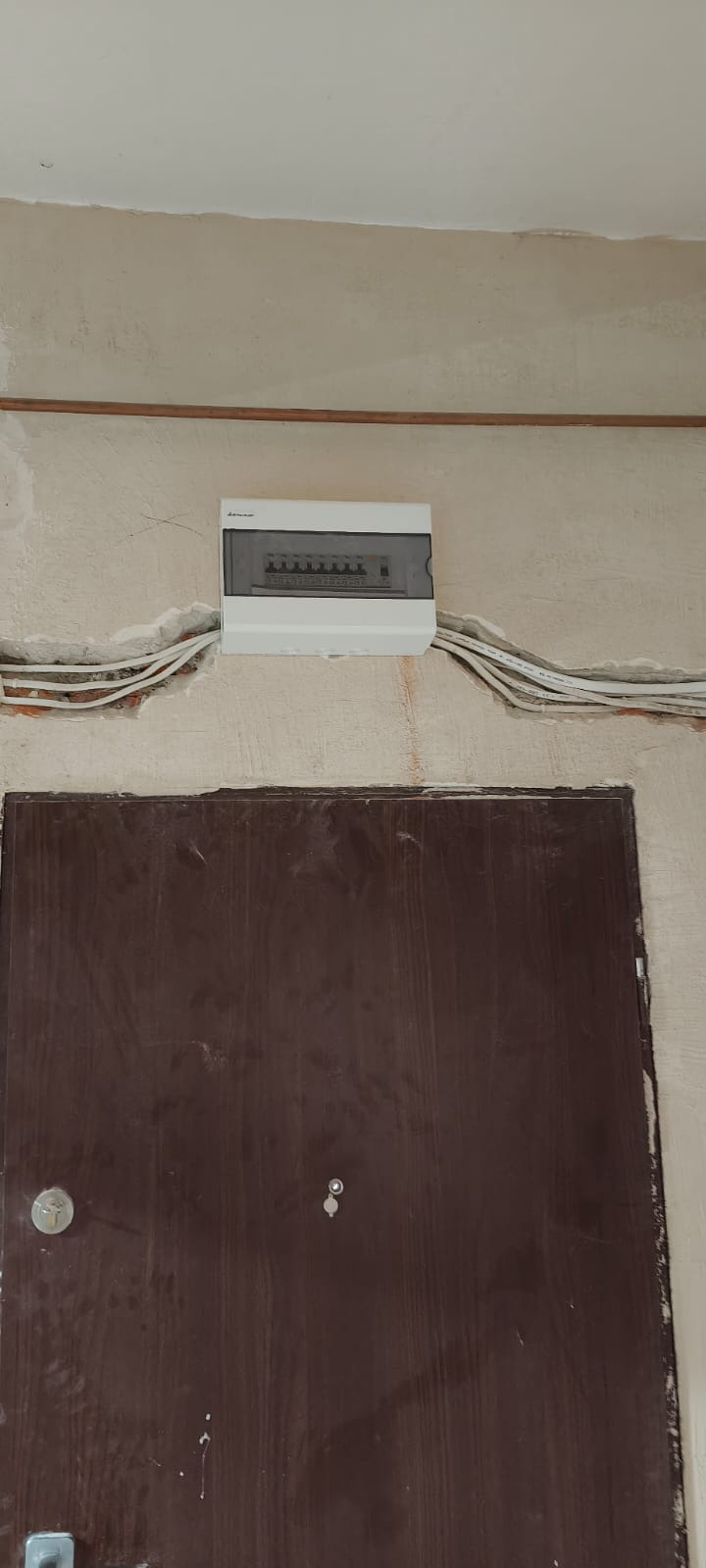 Biała skrzynka elektryczna z przewodami na ścianie nad brązowymi drzwiami. Ściana w stanie surowym, przygotowana do dalszych prac wykończeniowych.