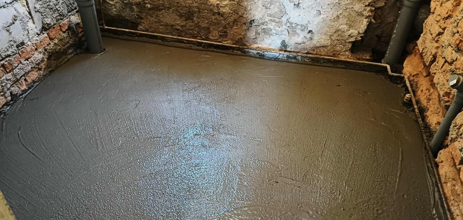 Świeżo wylana, mokra wylewka podłogowa w pomieszczeniu z surowymi ścianami z cegły i kamienia, widoczne rury instalacyjne przy ścianach.