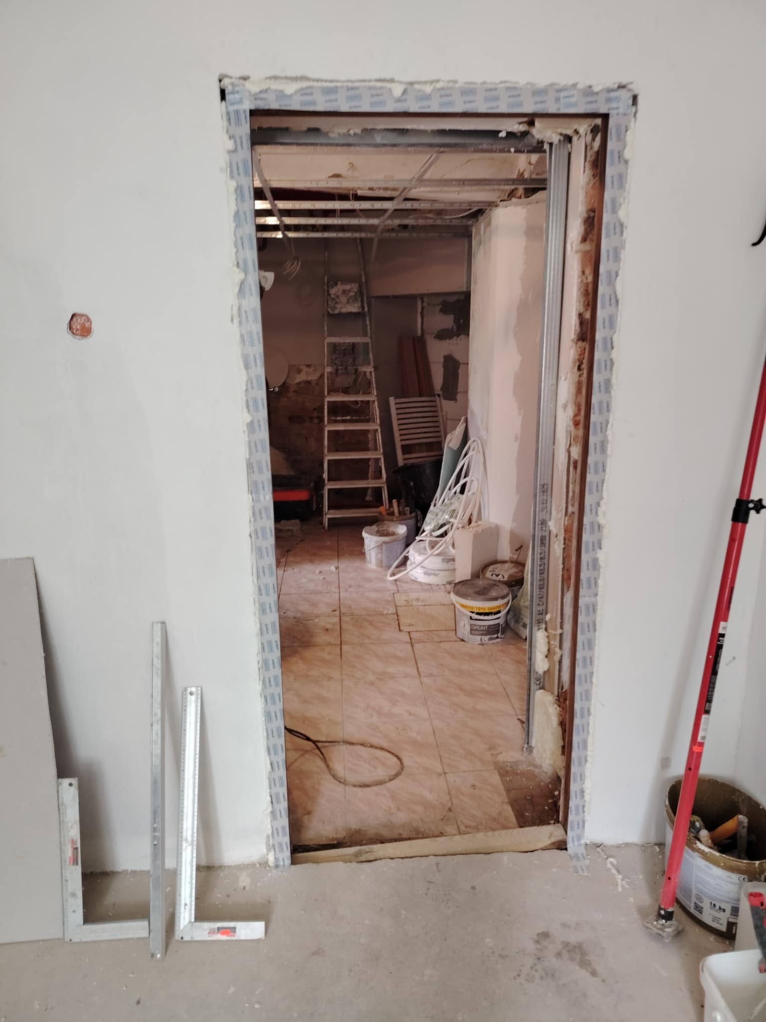 Przygotowanie do montażu ościeżnicy w surowym otworze drzwiowym. Widoczne profile, pianka montażowa, narzędzia i fragment pomieszczenia w trakcie prac wykończeniowych.