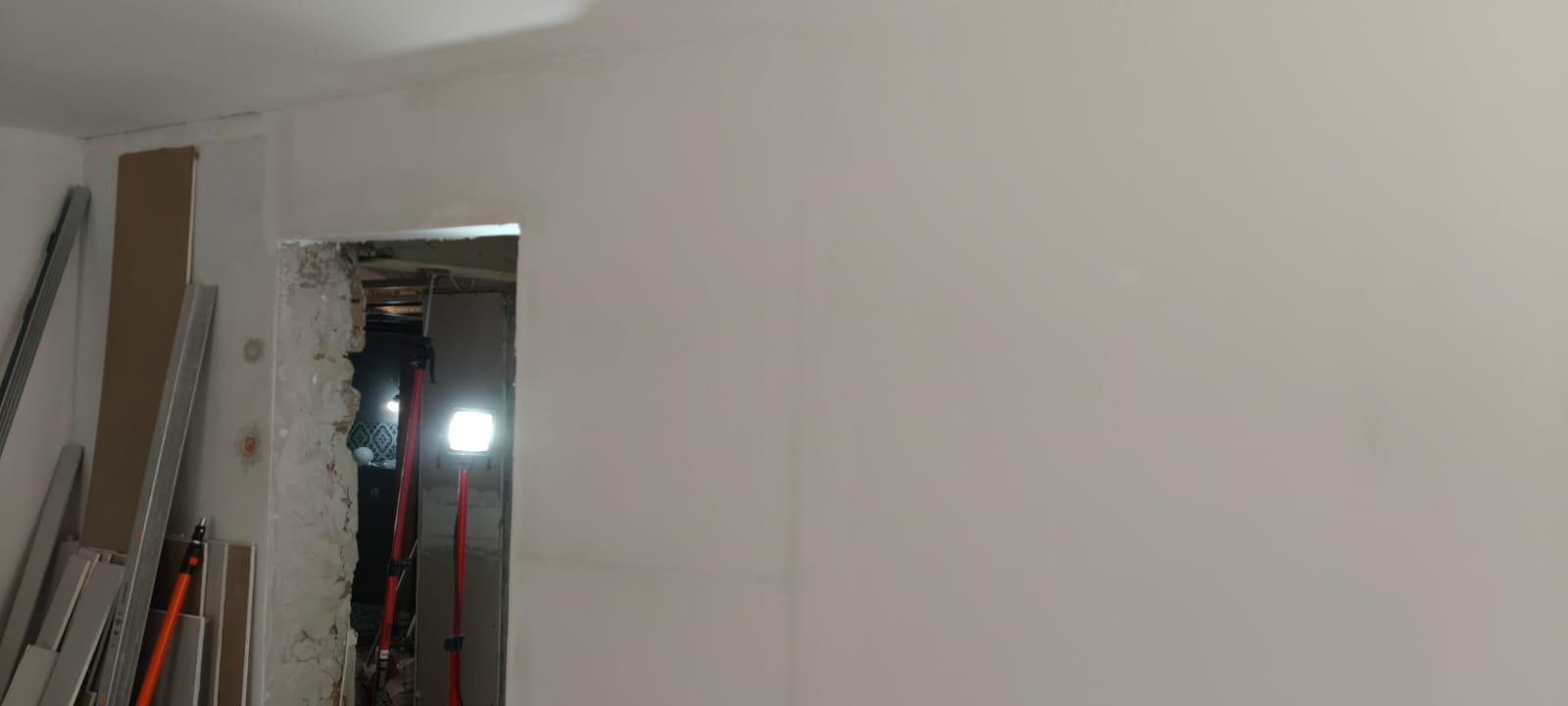 Wnętrze w trakcie prac wykończeniowych: gładkie, pomalowane ściany kontrastują z surowym otworem drzwiowym. Widoczne płyty karton-gips i oświetlenie budowlane.