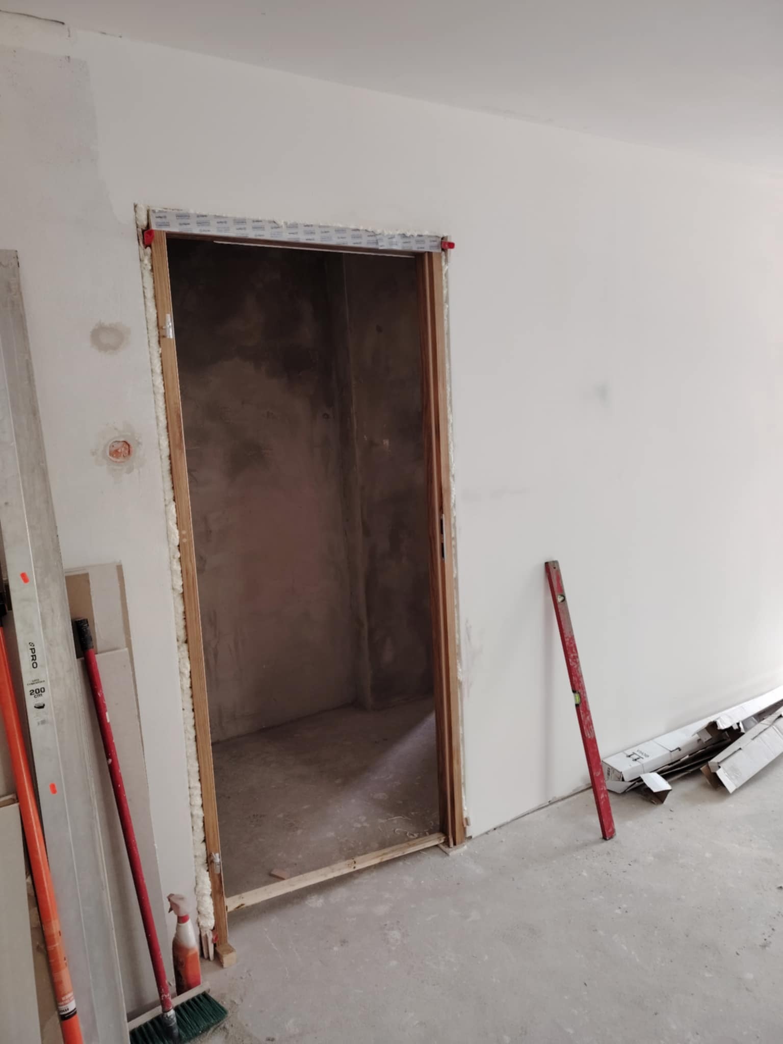 Przygotowany otwór drzwiowy z ościeżnicą, wypełniony pianką montażową, poziomica oparta o ścianę, widoczne narzędzia i materiały budowlane. Surowy stan wykończenia.