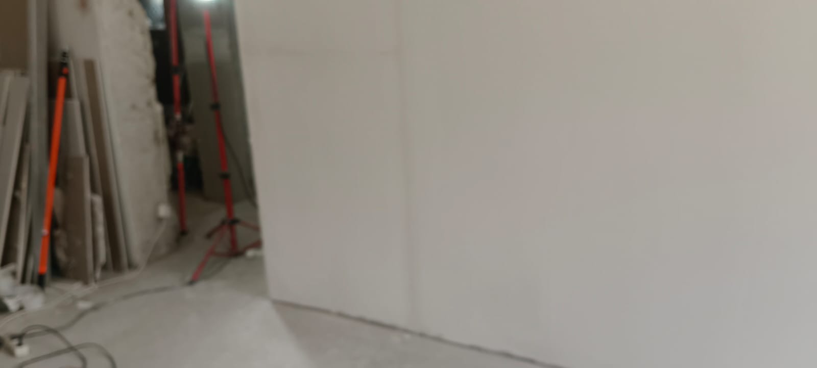 Ściana po szpachlowaniu i malowaniu na gładko, widoczne narzędzia i materiały budowlane w tle. Oświetlenie budowlane. Prace wykończeniowe w trakcie.