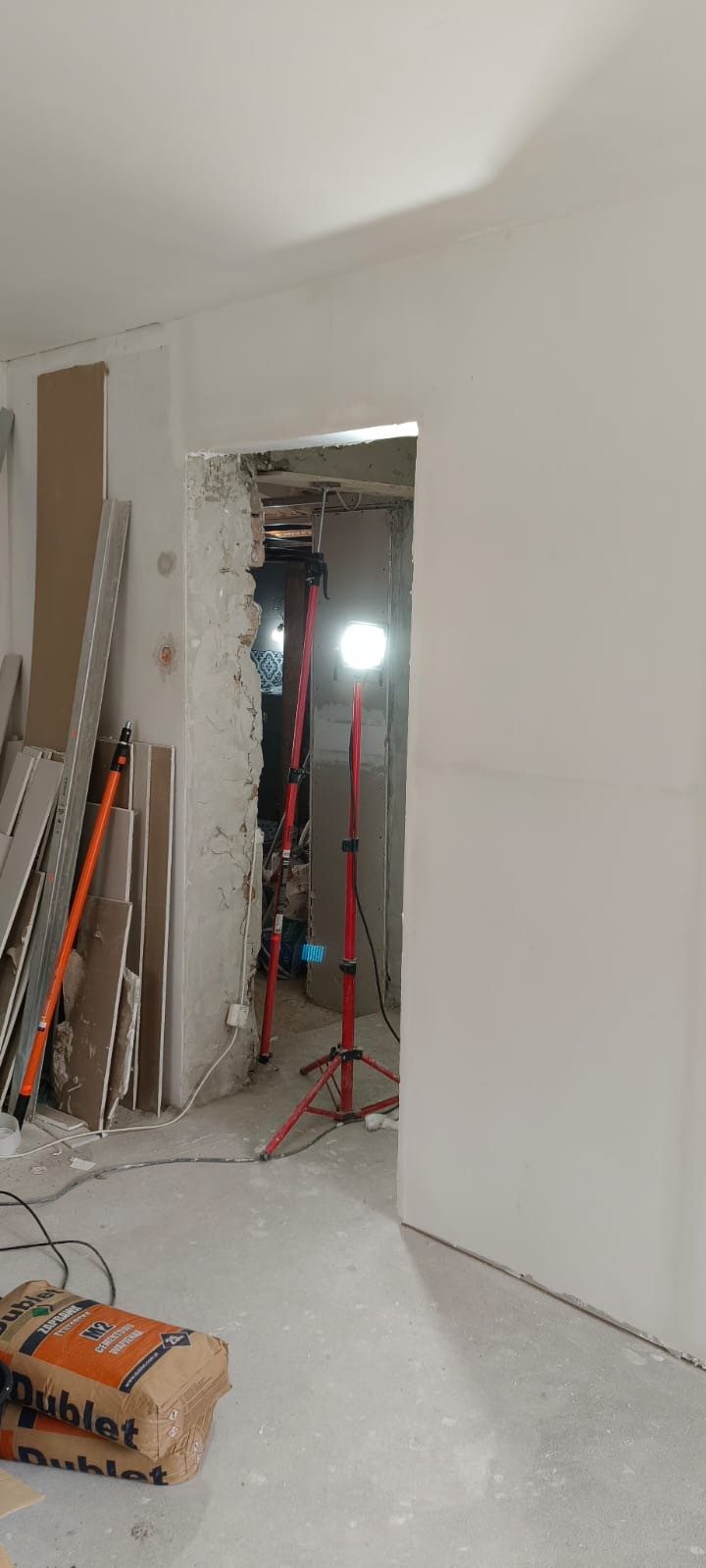 Wnętrze w trakcie remontu: oświetlona lampami ściana po szpachlowaniu, widoczne płyty karton-gips i worki z zaprawą. Przejście w stanie surowym.