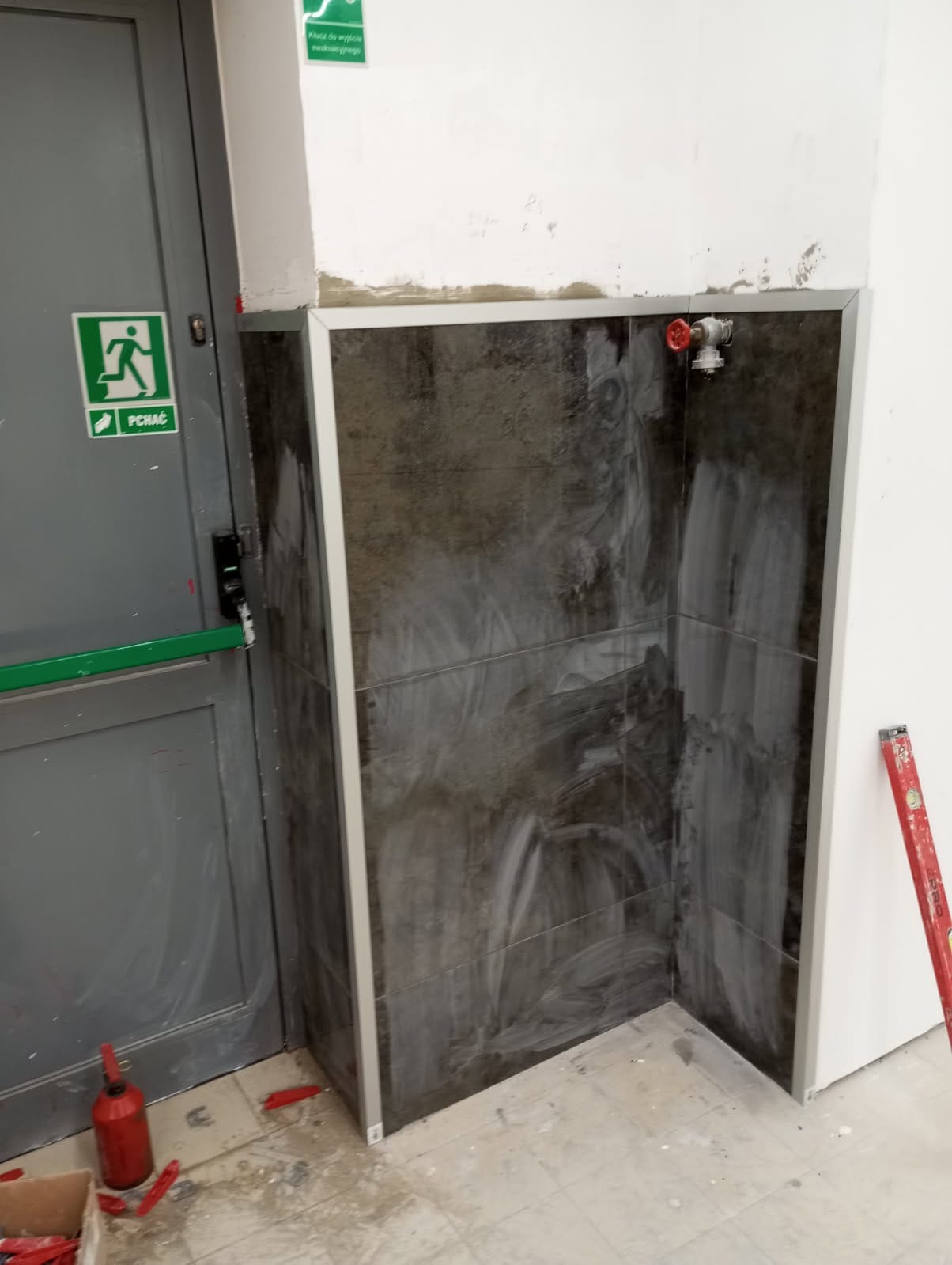 Ciemne płytki imitujące kamień, ułożone w rogu pomieszczenia, z widocznymi śladami fugowania i czerwoną armaturą. Poziomica oparta o ścianę, drzwi ewakuacyjne w tle.