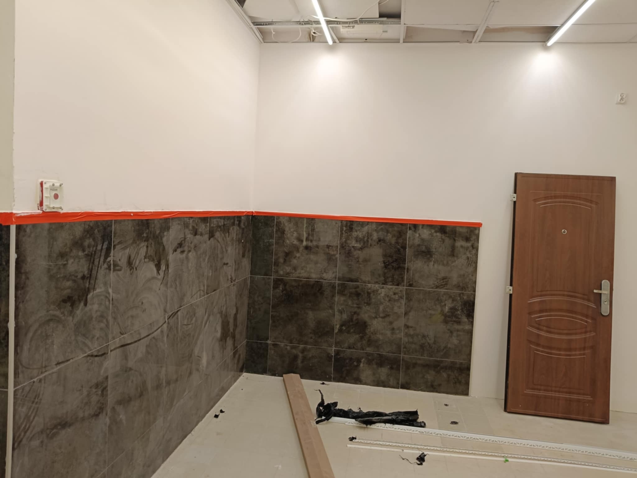 Wnętrze w trakcie prac wykończeniowych: częściowo położone ciemne płytki na ścianie, zabezpieczone pomarańczową taśmą, sufit podwieszany, drzwi wejściowe.