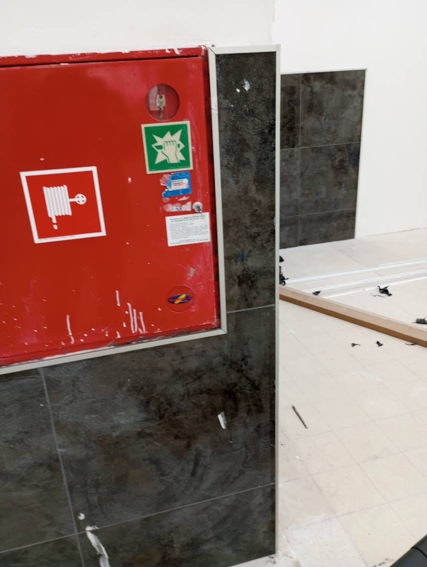 Wykończona ściana z ciemnymi płytkami, obok czerwona szafka hydrantowa. Widoczne ślady po remoncie na podłodze. Ukończone prace glazurnicze w obiekcie.