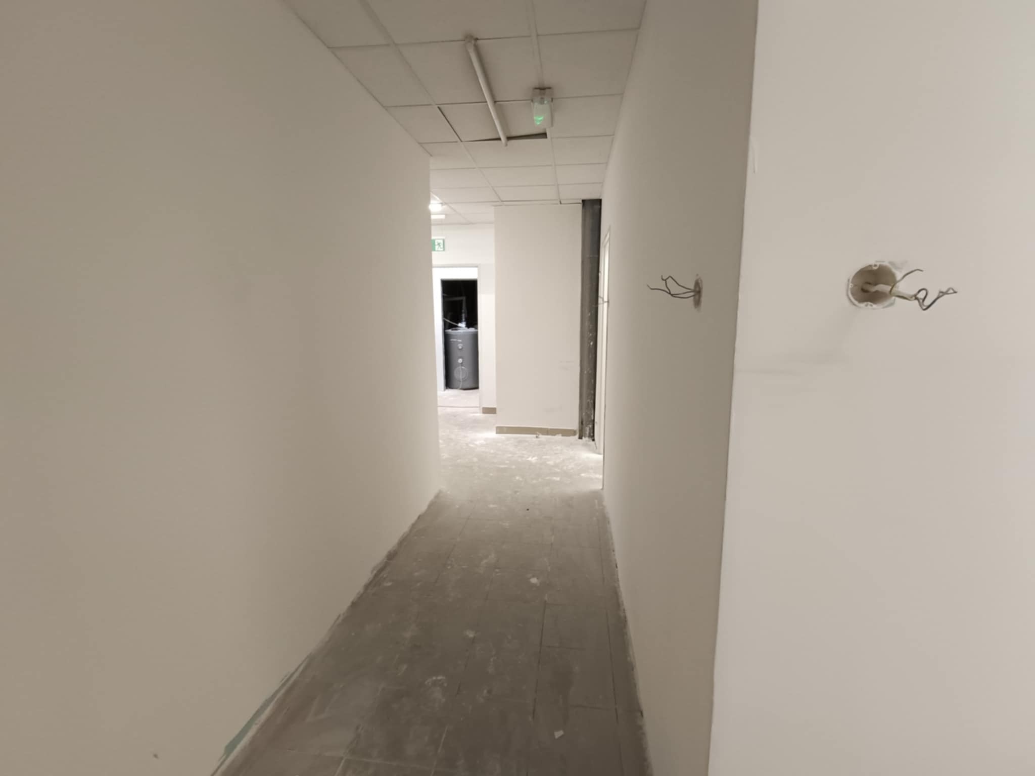 Korytarz w trakcie wykańczania: świeżo pomalowane ściany na biało, widoczne puszki elektryczne bez osprzętu, sufit z kasetonów. Podłoga częściowo zakryta folią.
