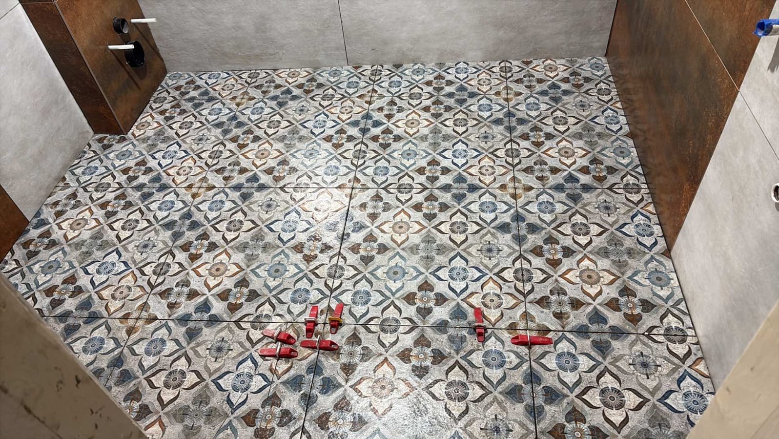 Ułożona podłoga z płytek w kwiatowy wzór, z czerwonymi klipsami poziomującymi. Widoczne fragmenty ściany z brązowymi i szarymi płytkami.