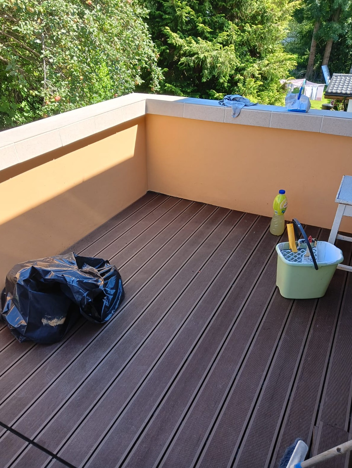 Balkon z kompozytowymi deskami podłogowymi w trakcie sprzątania. Widoczny worek na śmieci, wiadro z narzędziami i butelka płynu czyszczącego. Słoneczny dzień.