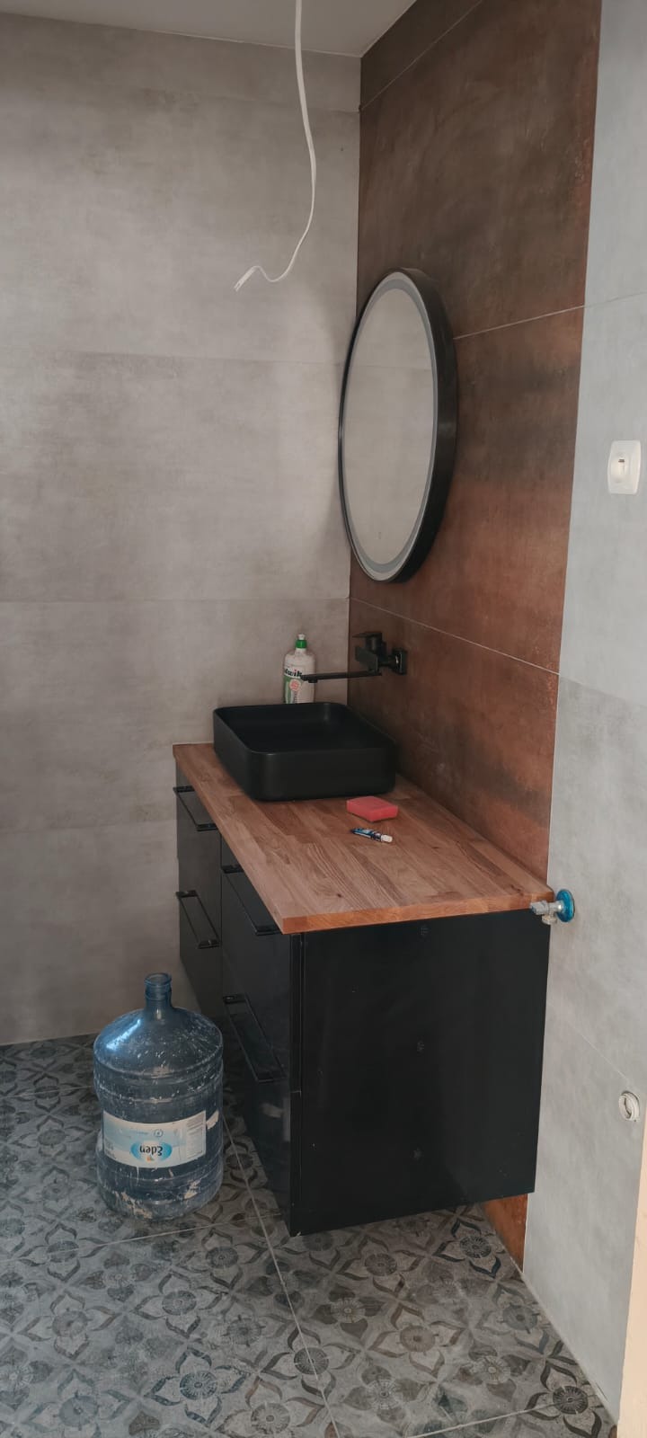 Nowoczesna łazienka po remoncie: czarna umywalka nablatowa, drewniany blat, okrągłe lustro z podświetleniem i dekoracyjne płytki ścienne w odcieniach szarości i brązu.