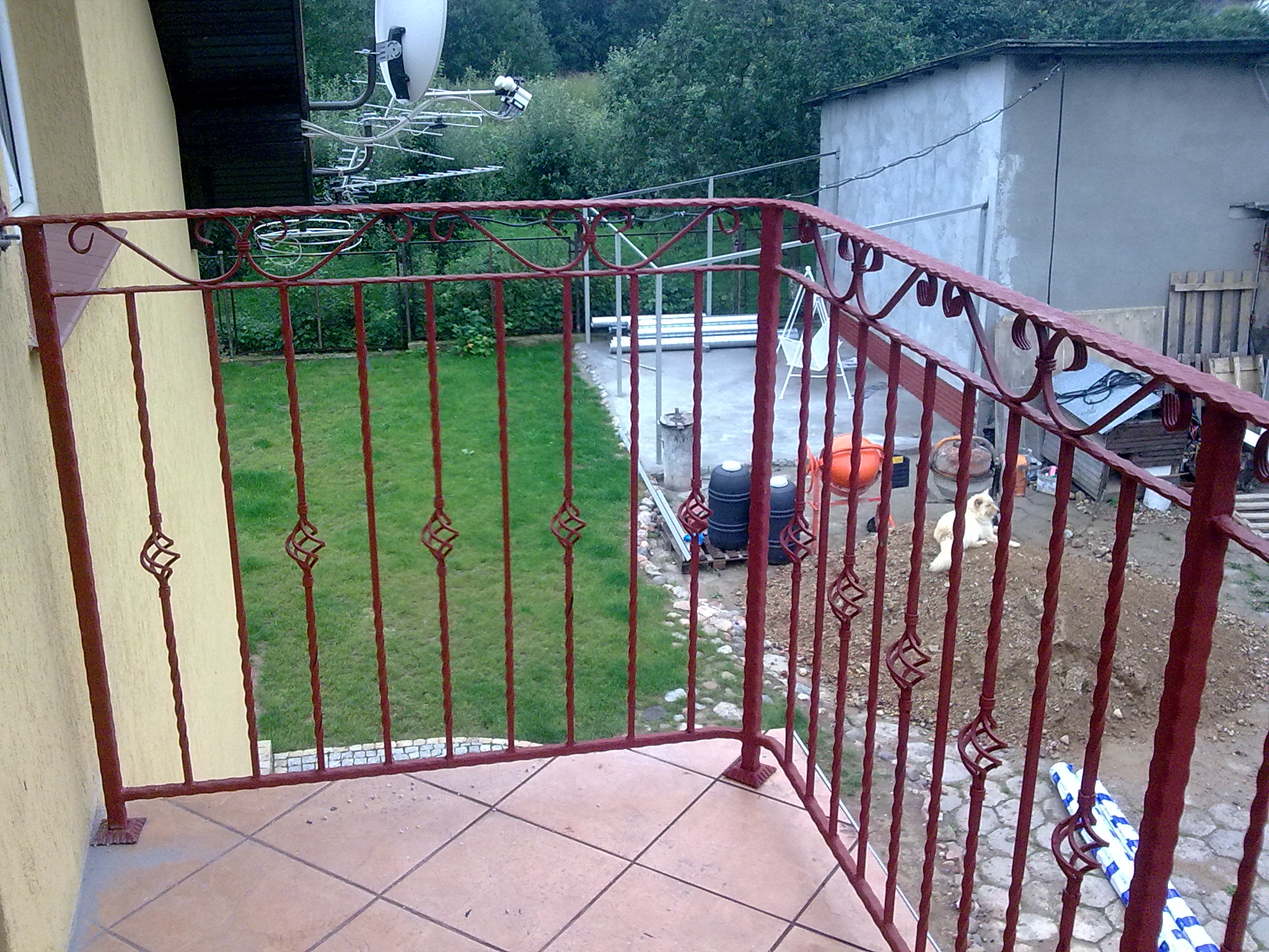 Czerwona metalowa balustrada balkonowa z ozdobnymi elementami w kształcie rombów, widok na trawnik i posesję z budynkiem gospodarczym w tle.