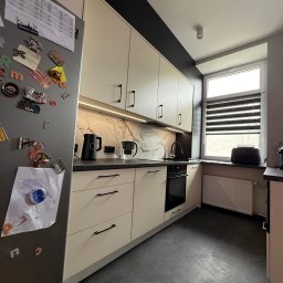 Artur Mlynarczyk - Jasna kuchnia na wymiar z czarnymi akcentami: blat, uchwyty, pas nad szafkami. Na lodówce rysunek dziecka i magnesy. Nowoczesne oświetlenie podszafkowe.