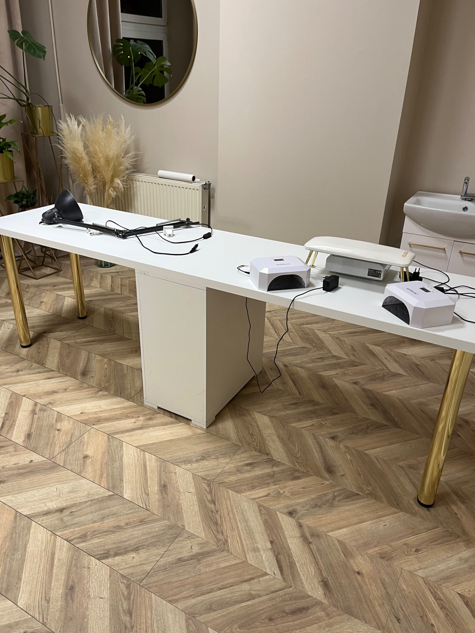 Długi, biały stół z lampą i urządzeniami do manicure, ustawiony w pomieszczeniu z parkietem w jodełkę i okrągłym lustrem na ścianie.