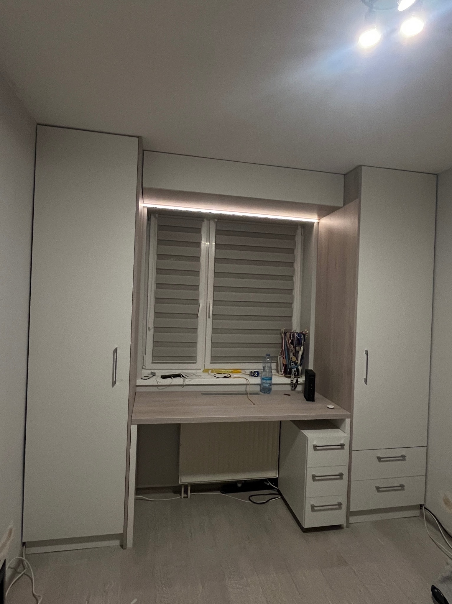 Biała szafa wnękowa z biurkiem pod oknem, oświetlenie LED nad blatem, szare rolety w oknie, minimalistyczny design, widok z przodu, jasne wnętrze.