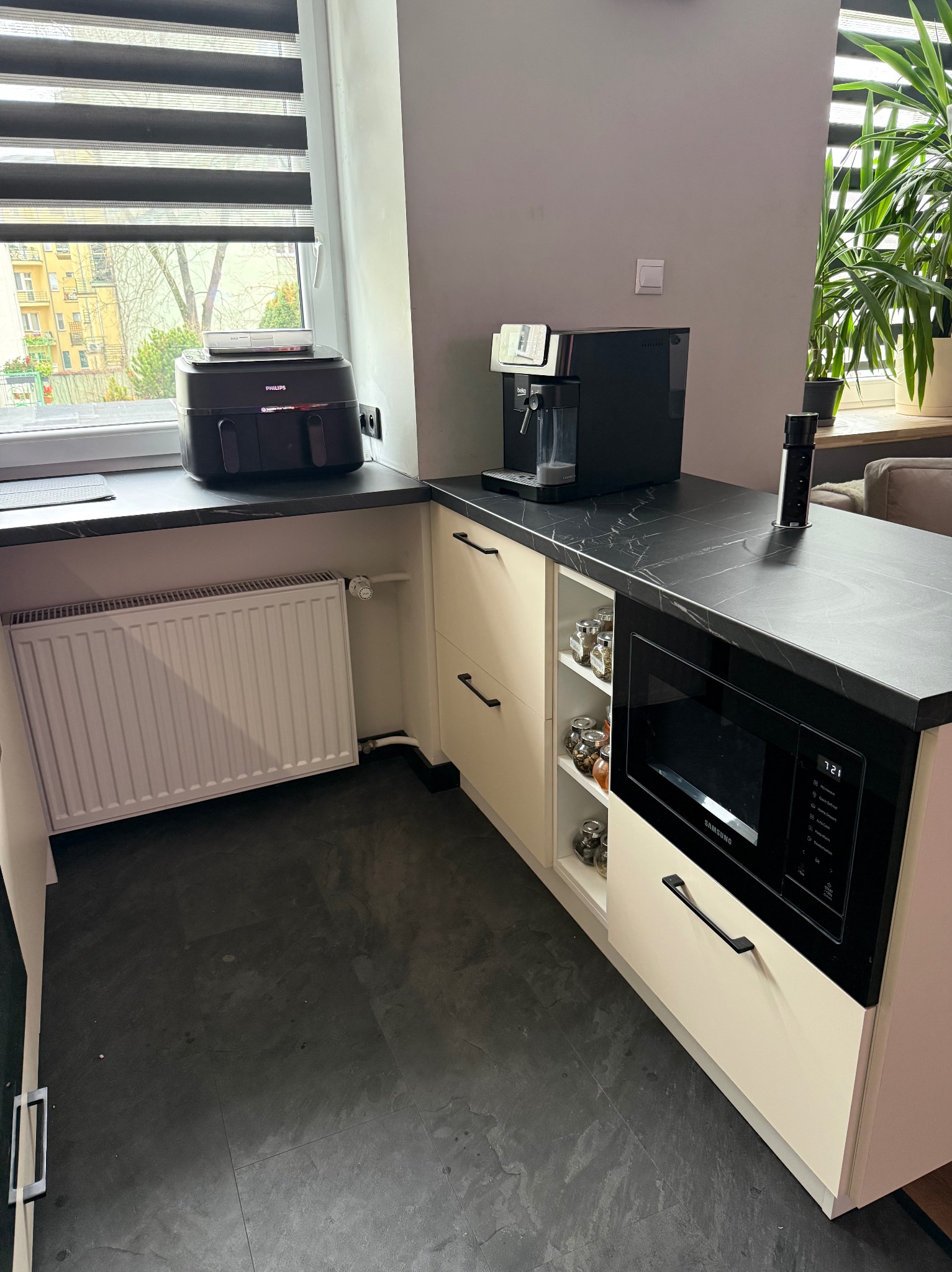 Jasna zabudowa kuchenna z czarnym blatem, na którym stoją ekspres do kawy i frytkownica. Widoczna mikrofalówka i przyprawy na półkach. Za oknem z roletami widok na budynki.