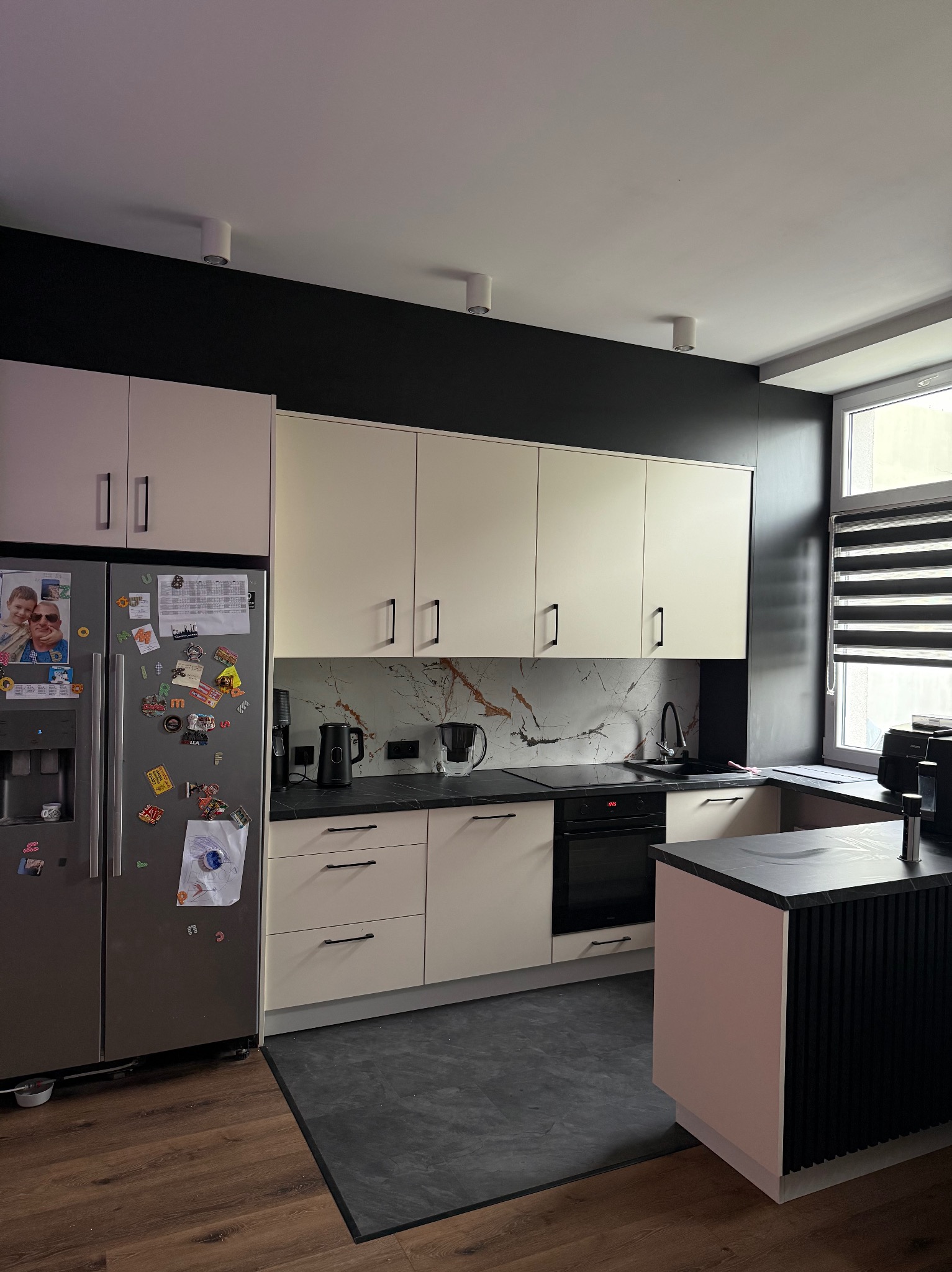 Jasna kuchnia z czarnymi akcentami: ściana, blat, dodatki. Lodówka z magnesami, drewniana podłoga i czarny dywanik. Nowoczesny design z marmurowym backsplash.