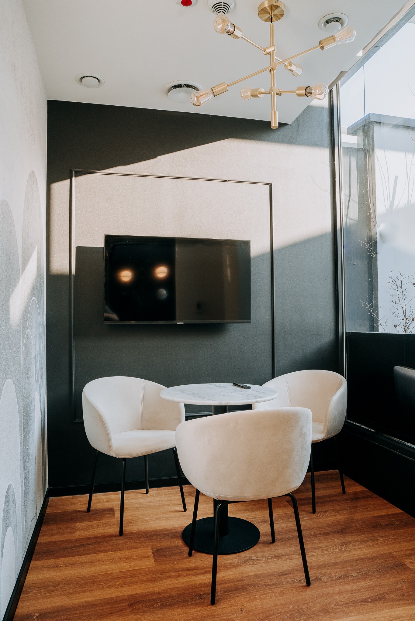 Nowoczesna sala lekcyjna z okrągłym stołem, trzema krzesłami i telewizorem na ścianie. Jasne wnętrze z drewnianą podłogą i designerską lampą sufitową.