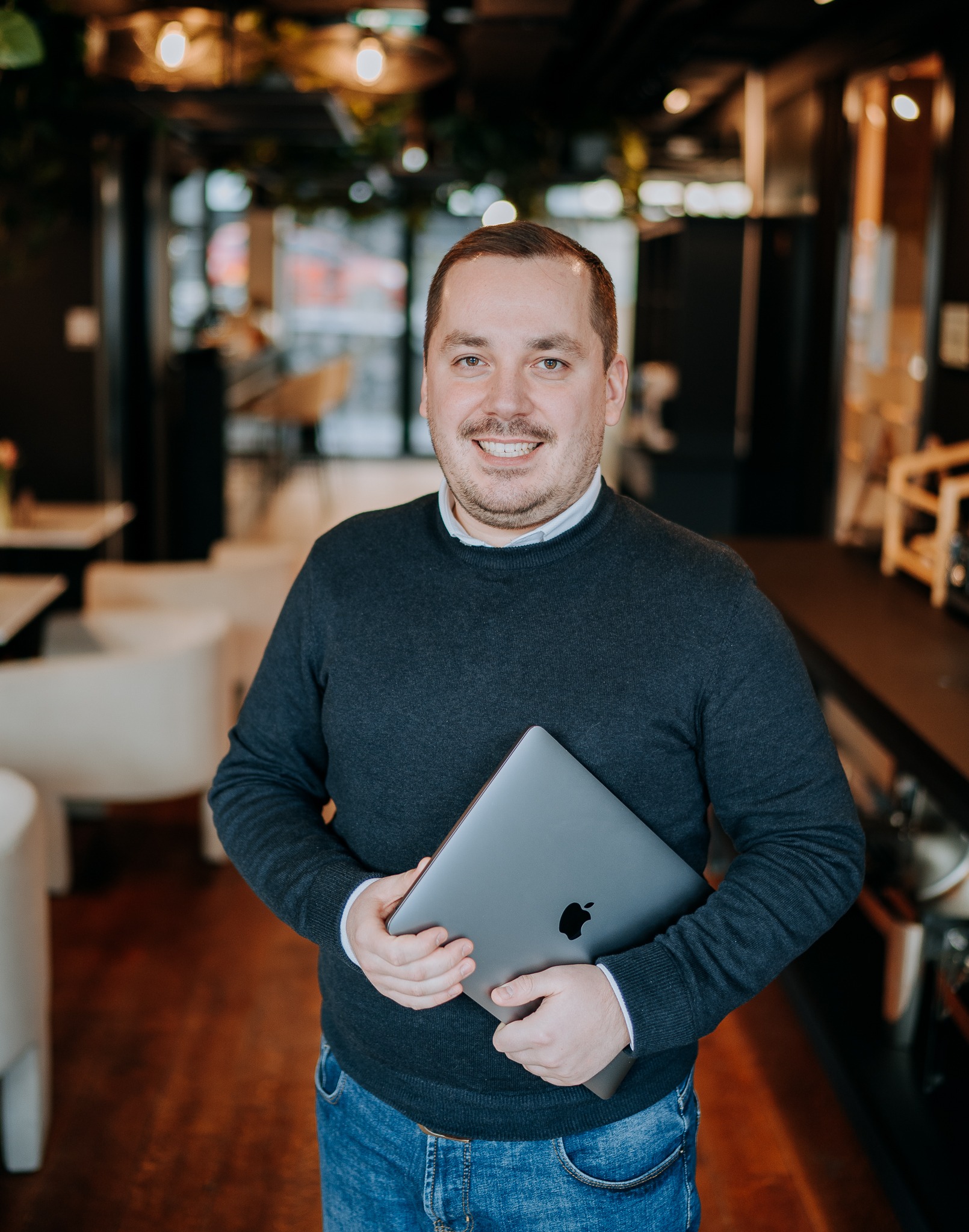 Uśmiechnięty lektor z laptopem Apple w dłoniach, w tle kawiarnia. Portret w swobodnej atmosferze, sugerujący nowoczesne metody nauczania.