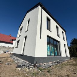 Zorganizowana Grupa Budowlana - Nowoczesny, biały dom z czarnymi akcentami, widoczny z dołu pod kątem. Elewacja świeżo po malowaniu, z widocznymi materiałami budowlanymi wokół. Błękitne niebo w tle.