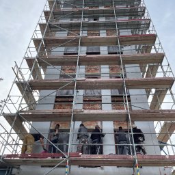 Zorganizowana Grupa Budowlana - Prace ociepleniowe na wysokim budynku w rusztowaniach. Widoczne fragmenty cegieł i szare płyty izolacyjne. Ekipa w trakcie pracy na różnych poziomach rusztowania.