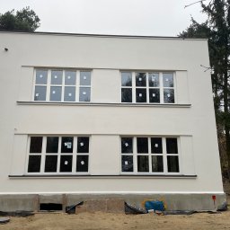 Zorganizowana Grupa Budowlana - Dwukondygnacyjny budynek po renowacji elewacji w stylu modernistycznym, z białymi ścianami i oknami z naklejkami, otoczony drzewami i piaskiem.