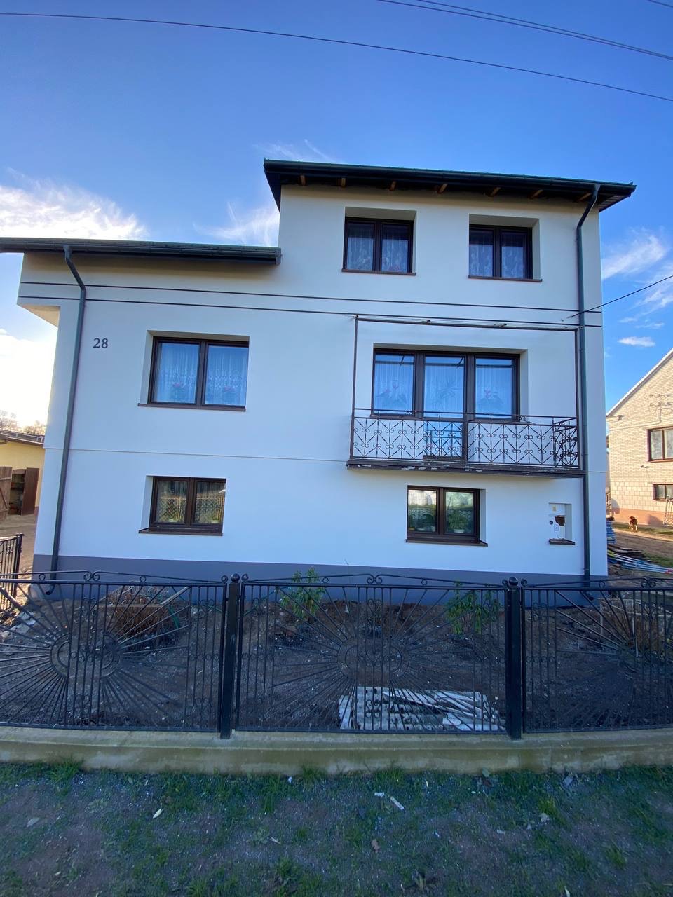 Dwupiętrowy dom z balkonem po renowacji elewacji, widoczny numer domu '28', ciemne okna, ozdobne metalowe ogrodzenie, błękitne niebo.