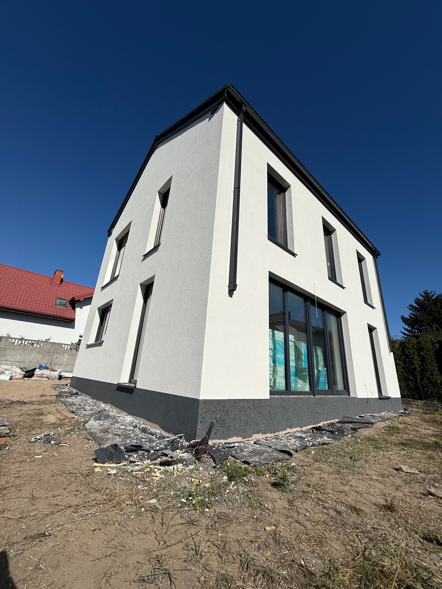 Nowoczesny, biały dom z czarnymi akcentami, widoczny z dołu pod kątem. Elewacja świeżo po malowaniu, z widocznymi materiałami budowlanymi wokół. Błękitne niebo w tle.