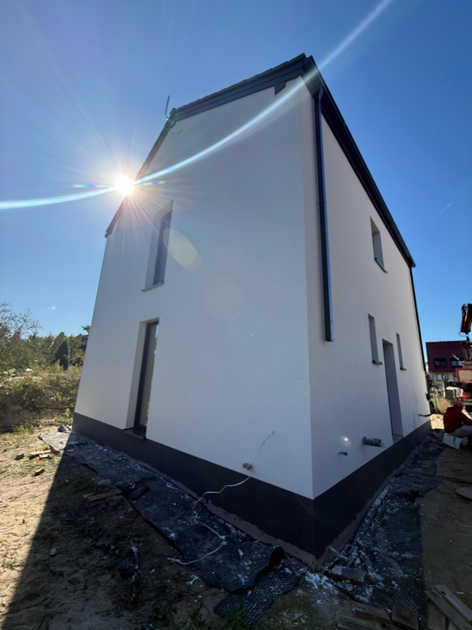 Biała elewacja domu jednorodzinnego w słoneczny dzień, widok z dołu, z czarnymi detalami wykończenia i widocznymi elementami przygotowania terenu.