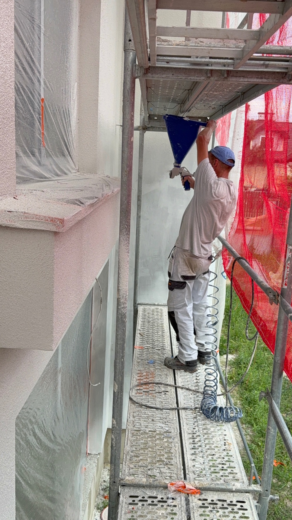 Pracownik na rusztowaniu aplikuje białą substancję na elewację budynku za pomocą specjalistycznego pistoletu natryskowego. Część elewacji zabezpieczona folią.