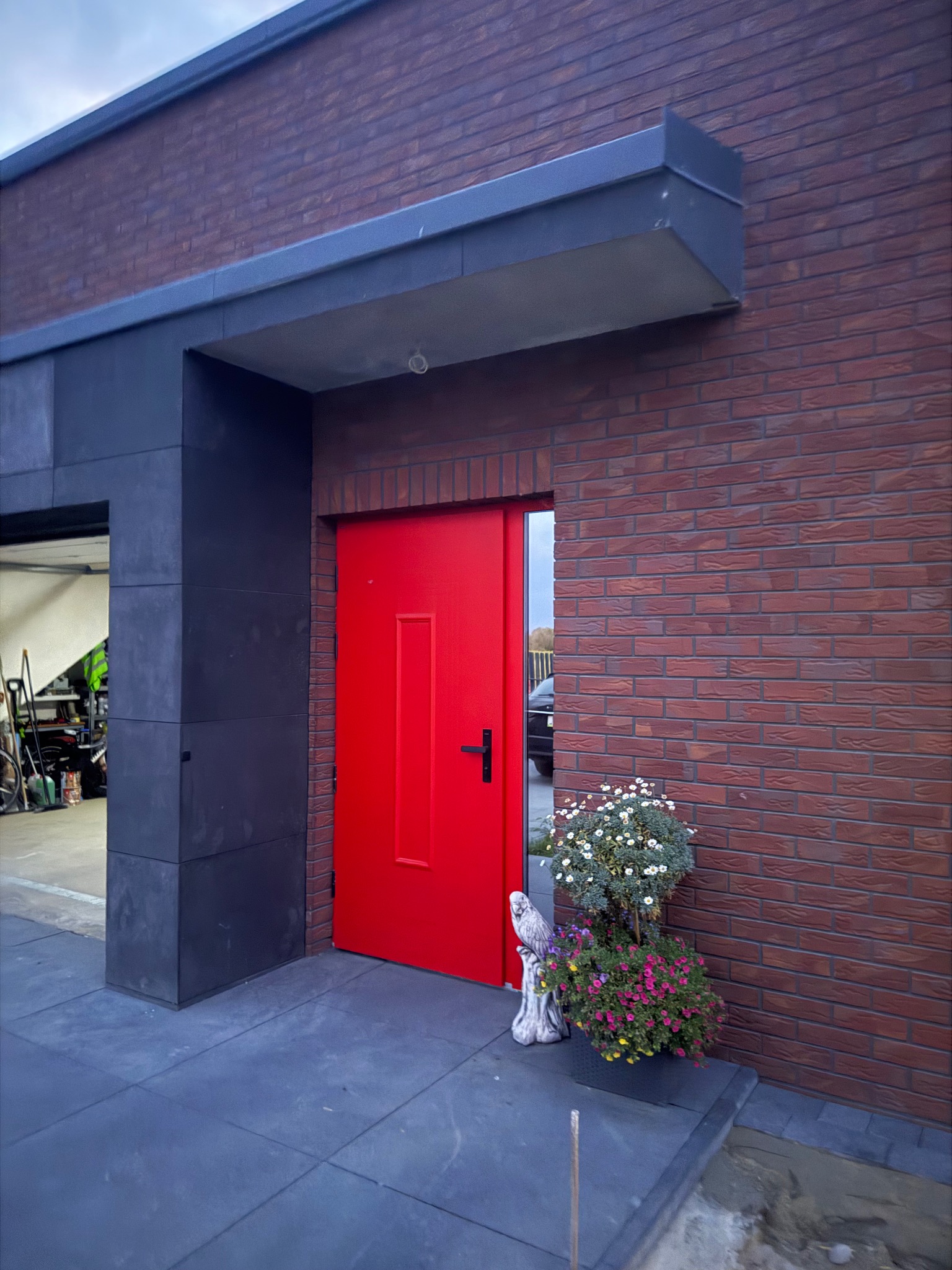 Ceglana elewacja z jaskrawoczerwonymi drzwiami wejściowymi, ozdobiona kwiatami i figurką sowy. Nowoczesne wykończenie z ciemnymi elementami.