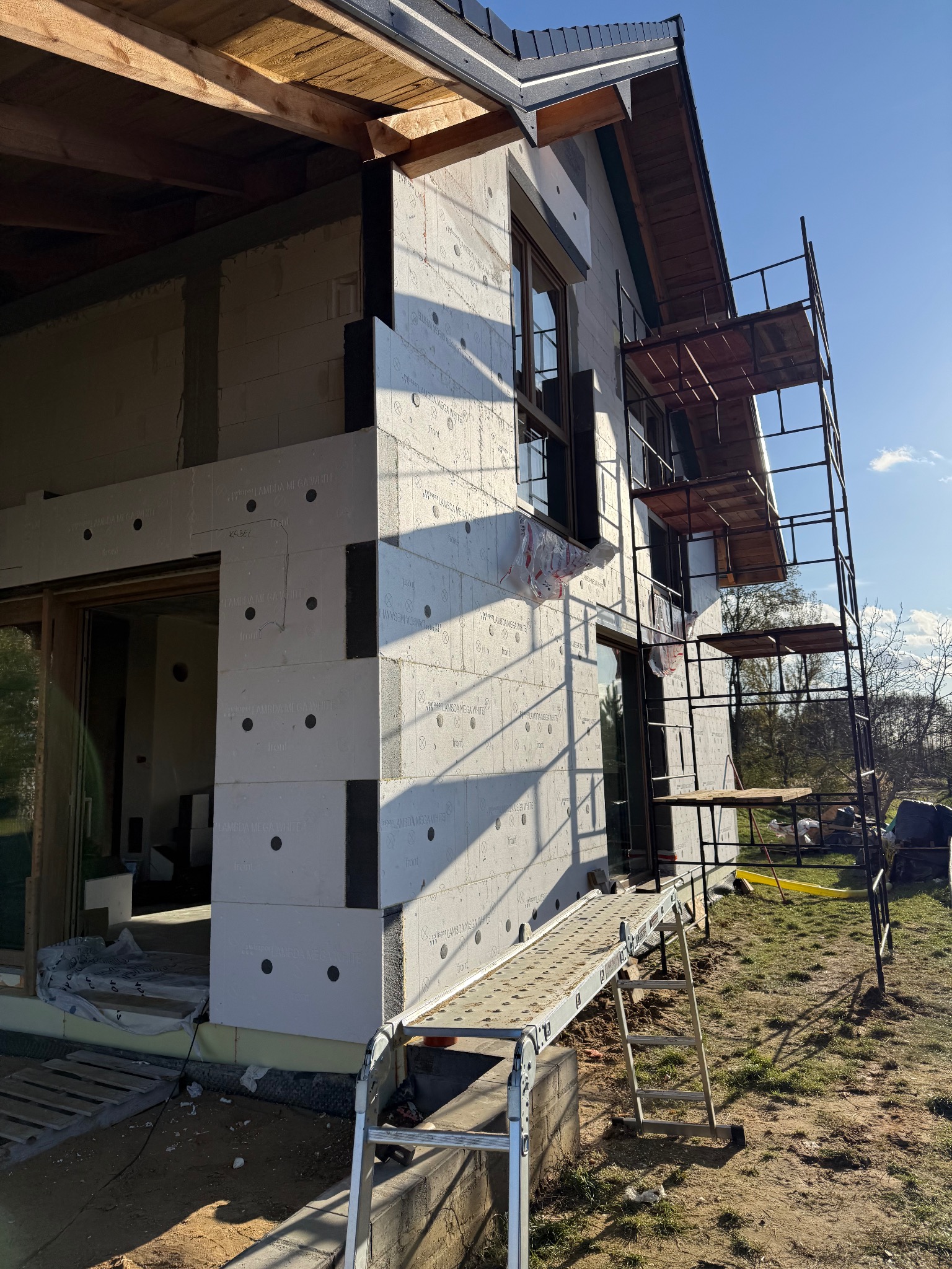 Dom w trakcie ocieplania: białe płyty izolacyjne z czarnymi punktami, rusztowanie przy ścianie, widoczne okna i fragment dachu. Słoneczny dzień.