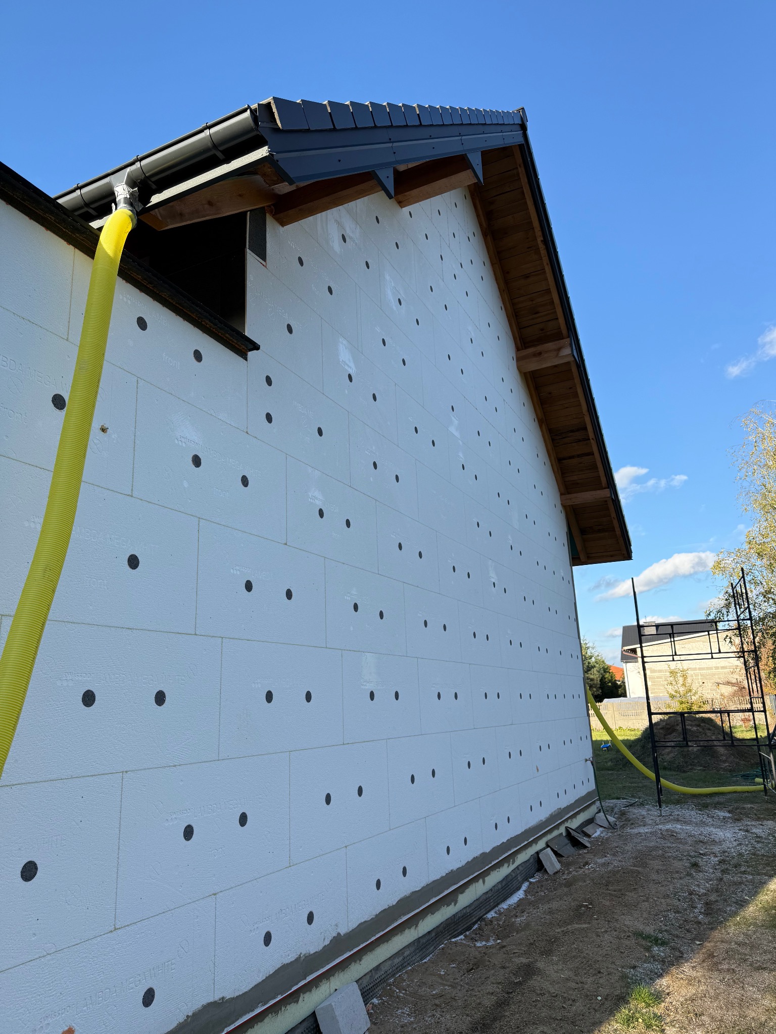 Białe płyty izolacyjne z czarnymi kołkami na ścianie budynku, żółty wąż doprowadzający materiał izolacyjny, widok z dołu na elewację w trakcie prac.