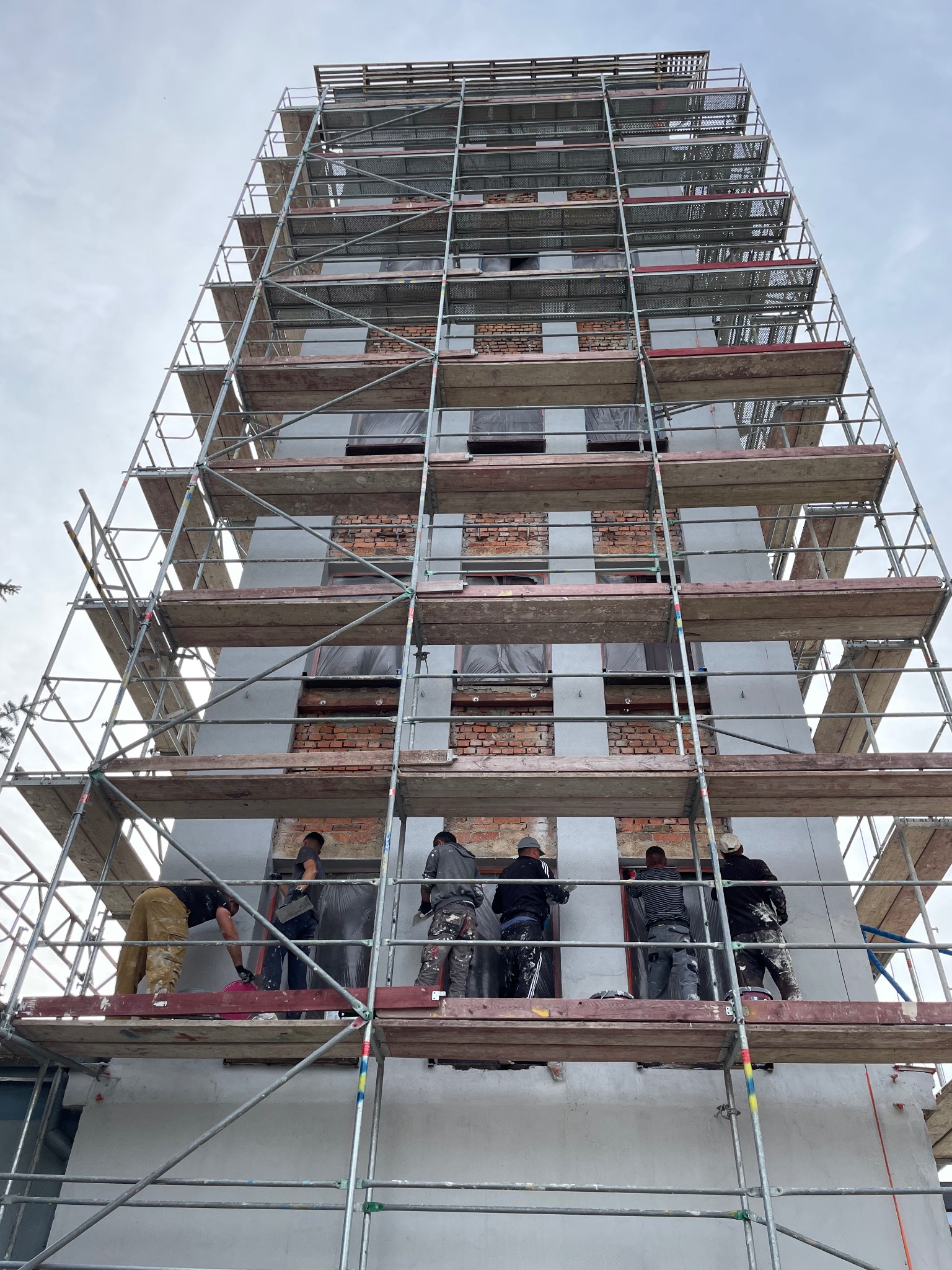 Prace ociepleniowe na wysokim budynku w rusztowaniach. Widoczne fragmenty cegieł i szare płyty izolacyjne. Ekipa w trakcie pracy na różnych poziomach rusztowania.