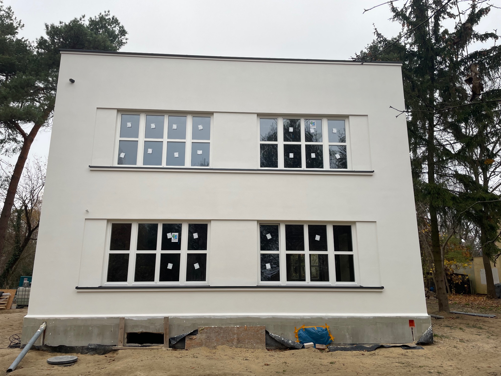 Dwukondygnacyjny budynek po renowacji elewacji w stylu modernistycznym, z białymi ścianami i oknami z naklejkami, otoczony drzewami i piaskiem.