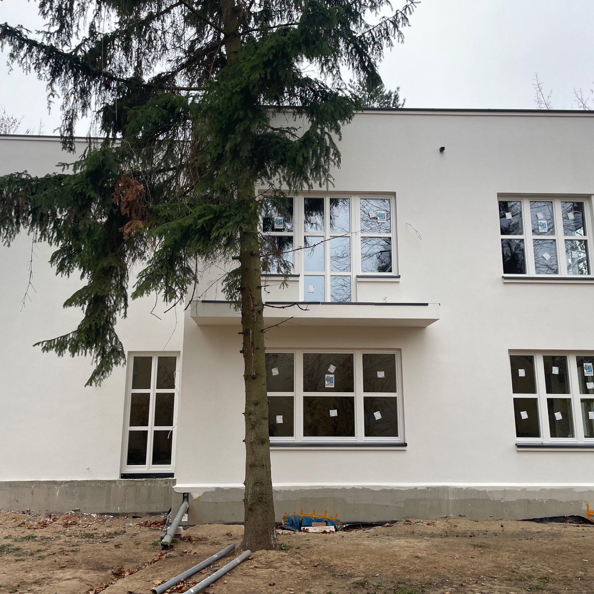 Biała elewacja budynku z nowymi oknami, częściowo zasłonięta przez drzewo. Widoczne rury na ziemi i przygotowany teren wokół budynku.