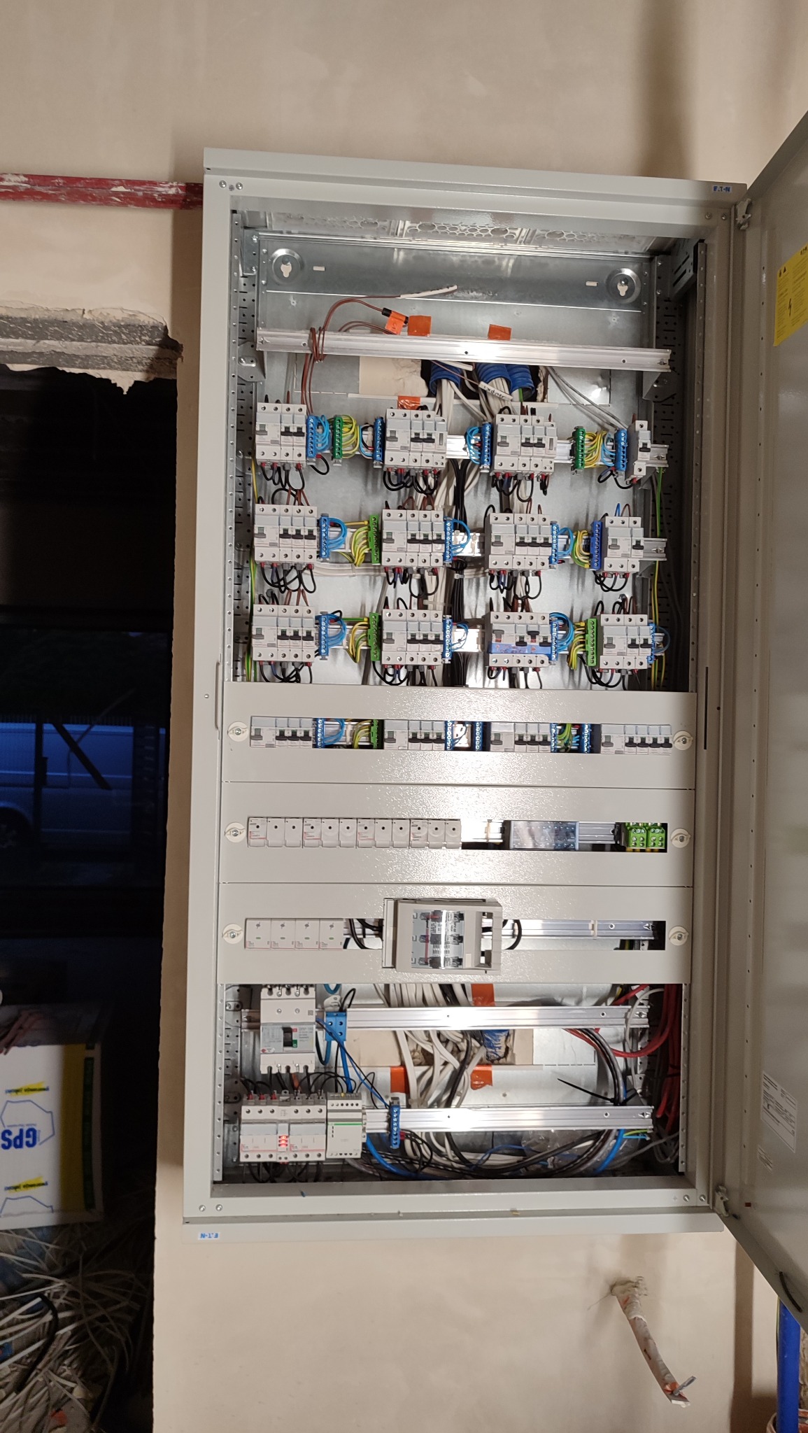 Otwarta skrzynka rozdzielcza z zainstalowanymi bezpiecznikami, przewodami i elementami elektrycznymi. Widoczne kable w różnych kolorach. Ściana w tle surowa, w trakcie wykończenia.