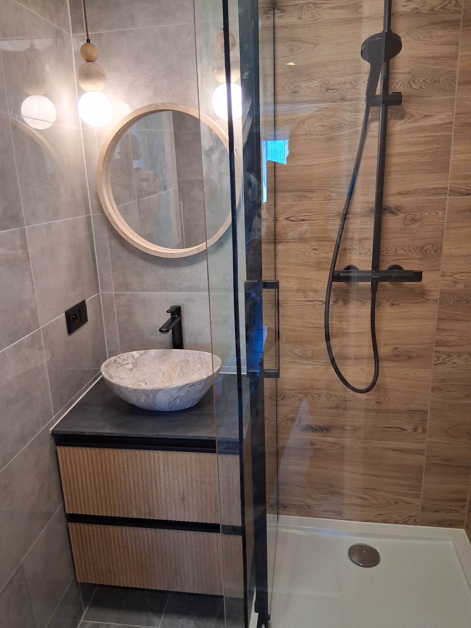 Nowoczesna łazienka z kabiną prysznicową typu walk-in, umywalką nablatową z marmuru i okrągłym lustrem. Ściany wyłożone płytkami imitującymi drewno i szarym gresem.