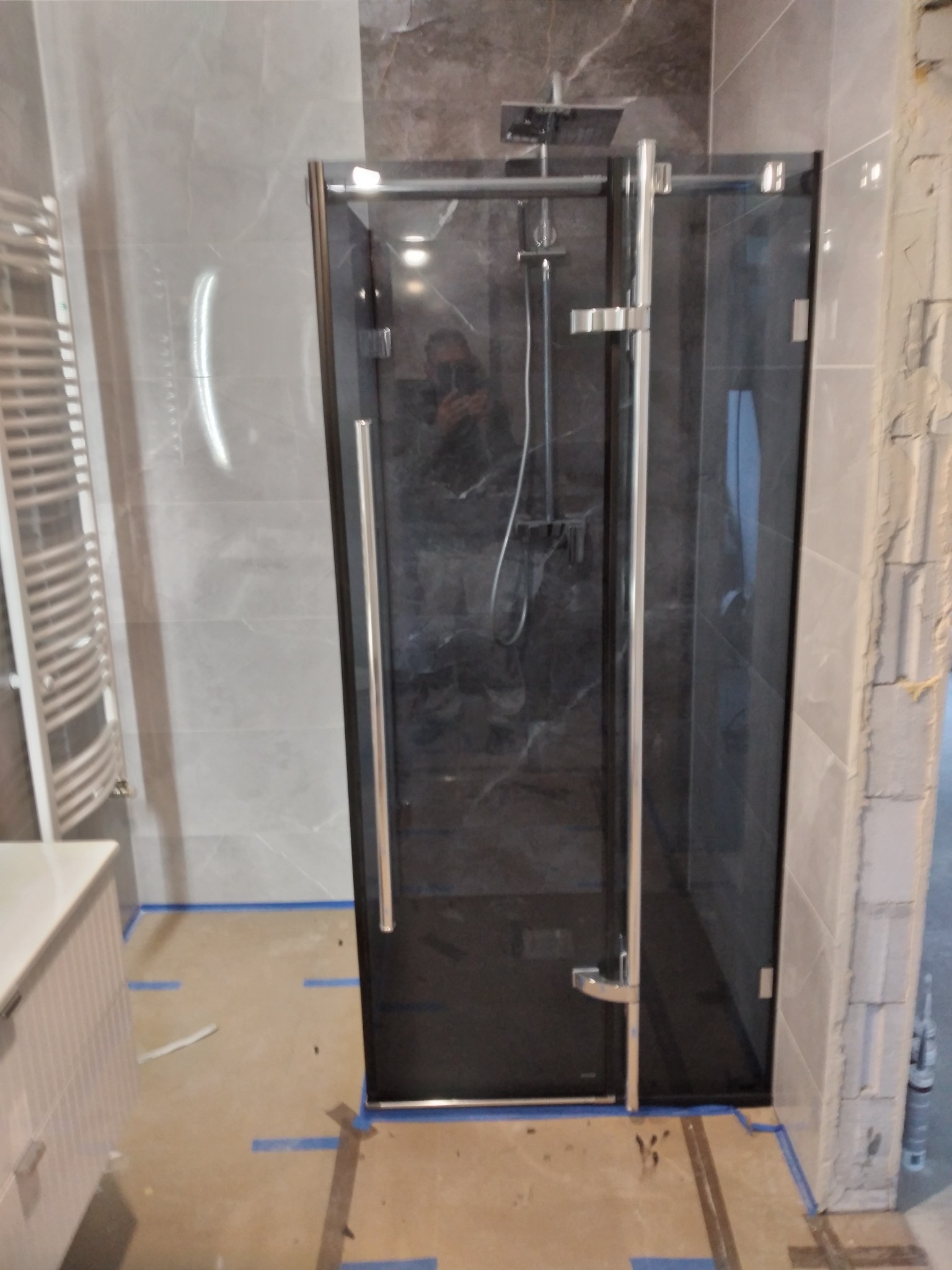 Nowoczesna kabina prysznicowa z ciemnymi płytkami w trakcie montażu, zabezpieczona taśmą malarską na podłodze. Widoczny fragment umywalki i grzejnika.