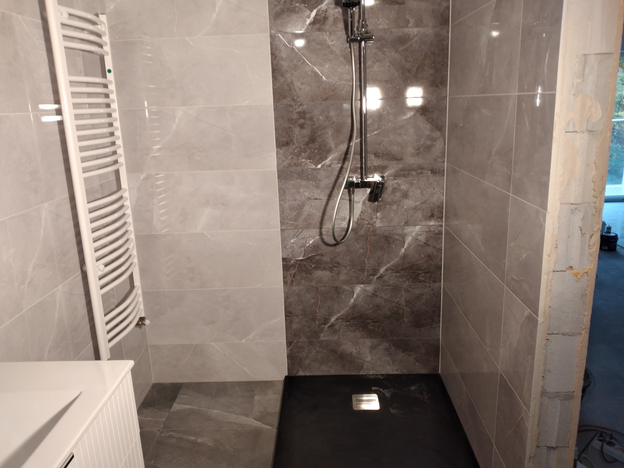Nowoczesna kabina prysznicowa z szarymi płytkami imitującymi marmur, czarnym brodzikiem i grzejnikiem drabinkowym. Widoczne niedokończone wykończenie ściany.