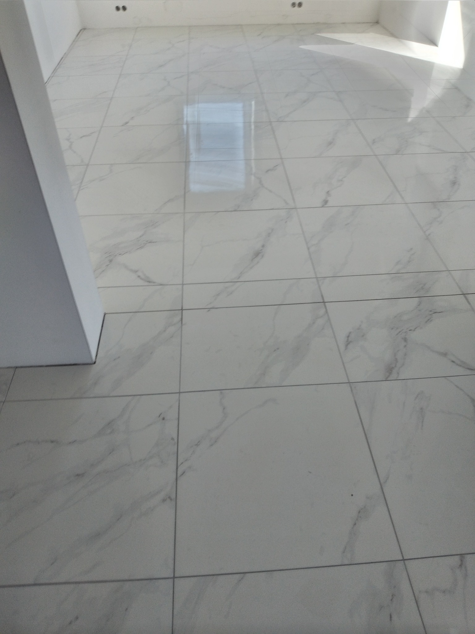 Białe, marmurowe płytki podłogowe w jasnym pomieszczeniu, ułożone równo z widocznymi fugami. Minimalistyczny design, świeżo po montażu, z odbiciem światła na powierzchni.