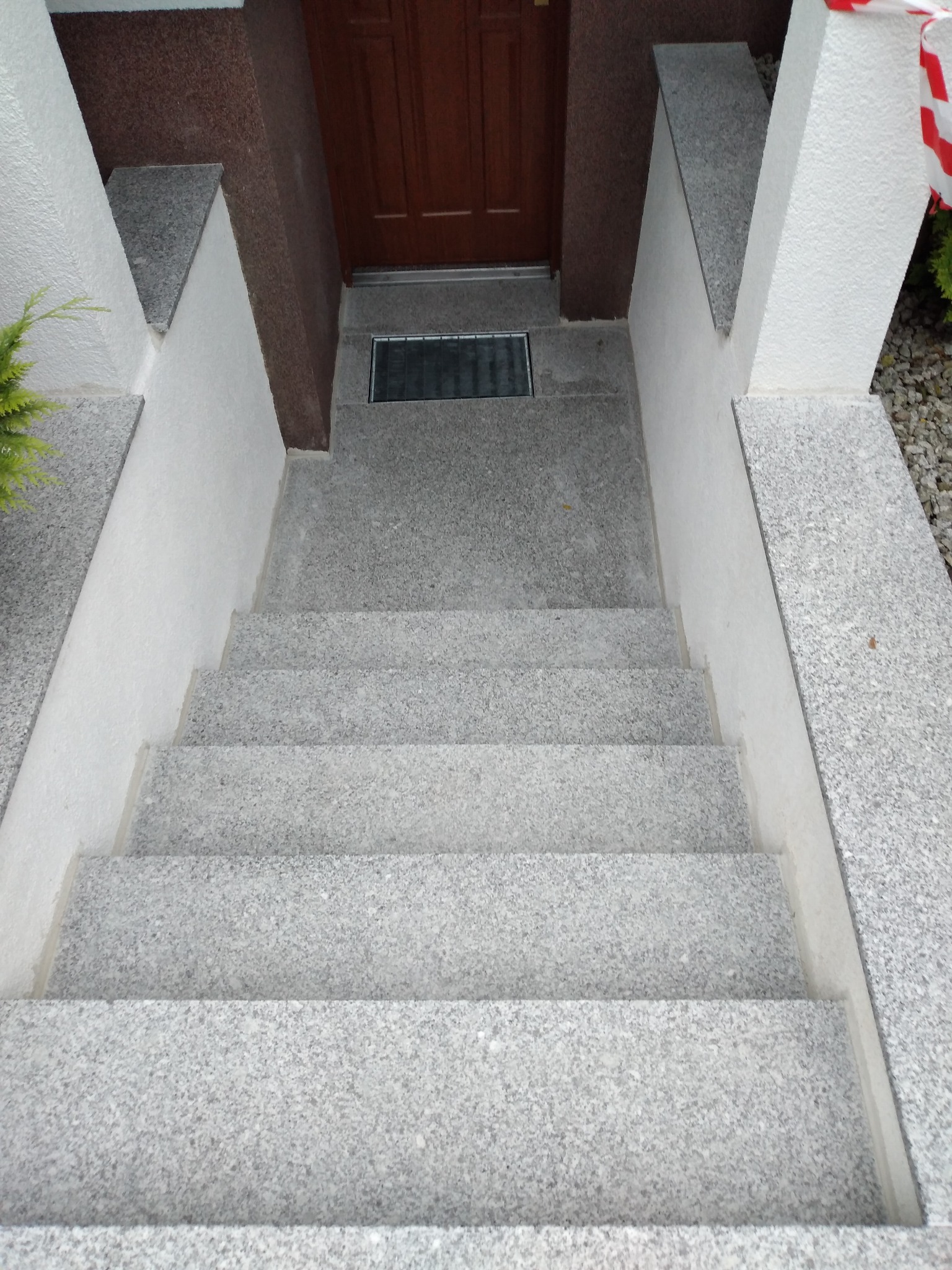 Granitowe schody wejściowe z brązowymi drzwiami w tle, wykończone białymi elementami. Widok z góry na szare stopnie i białe ściany.