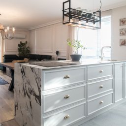 TREEO ART BARTŁOMIEJ STOKŁOSA, SEBASTIAN RZEPECKI S.C. - Jasna kuchnia z wyspą o marmurowym blacie i białymi szafkami. W tle salon z drewnianym stołem i kanapą. Nad wyspą wisi czarna, metalowa lampa.