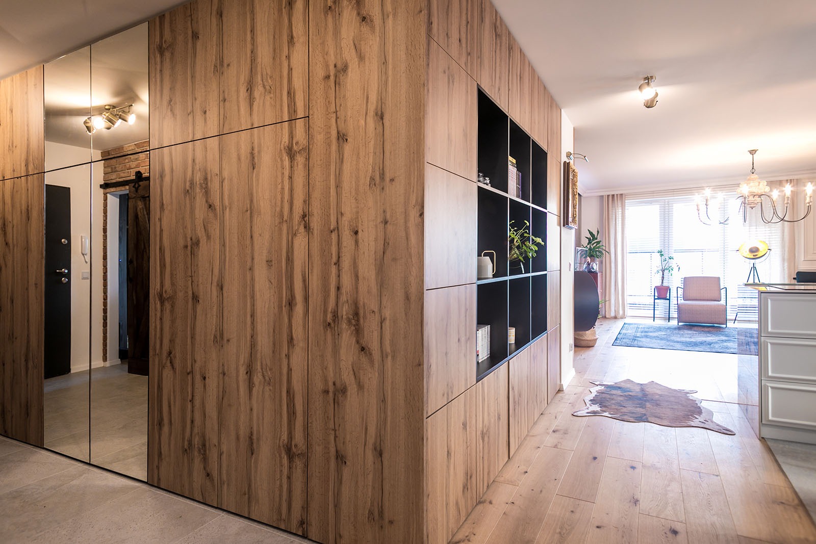 Drewniana szafa wnękowa z lustrzanymi drzwiami i otwartymi półkami, w jasnym wnętrzu z drewnianą podłogą i widokiem na salon.