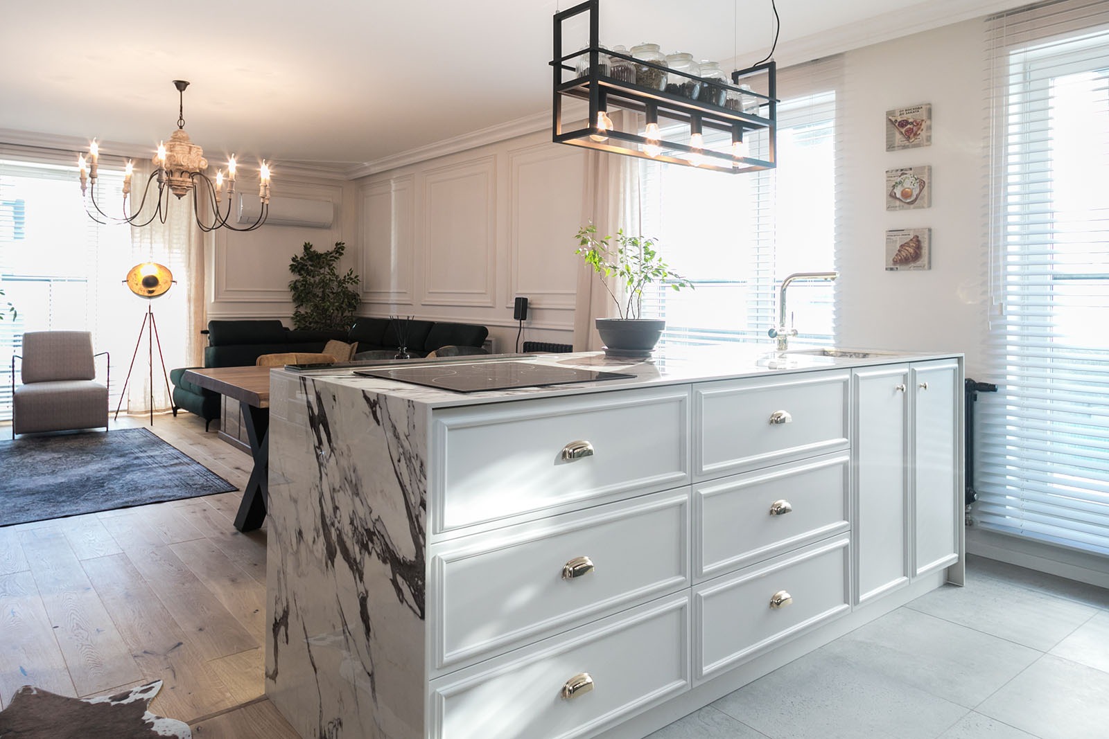 Jasna kuchnia z wyspą o marmurowym blacie i białymi szafkami. W tle salon z drewnianym stołem i kanapą. Nad wyspą wisi czarna, metalowa lampa.