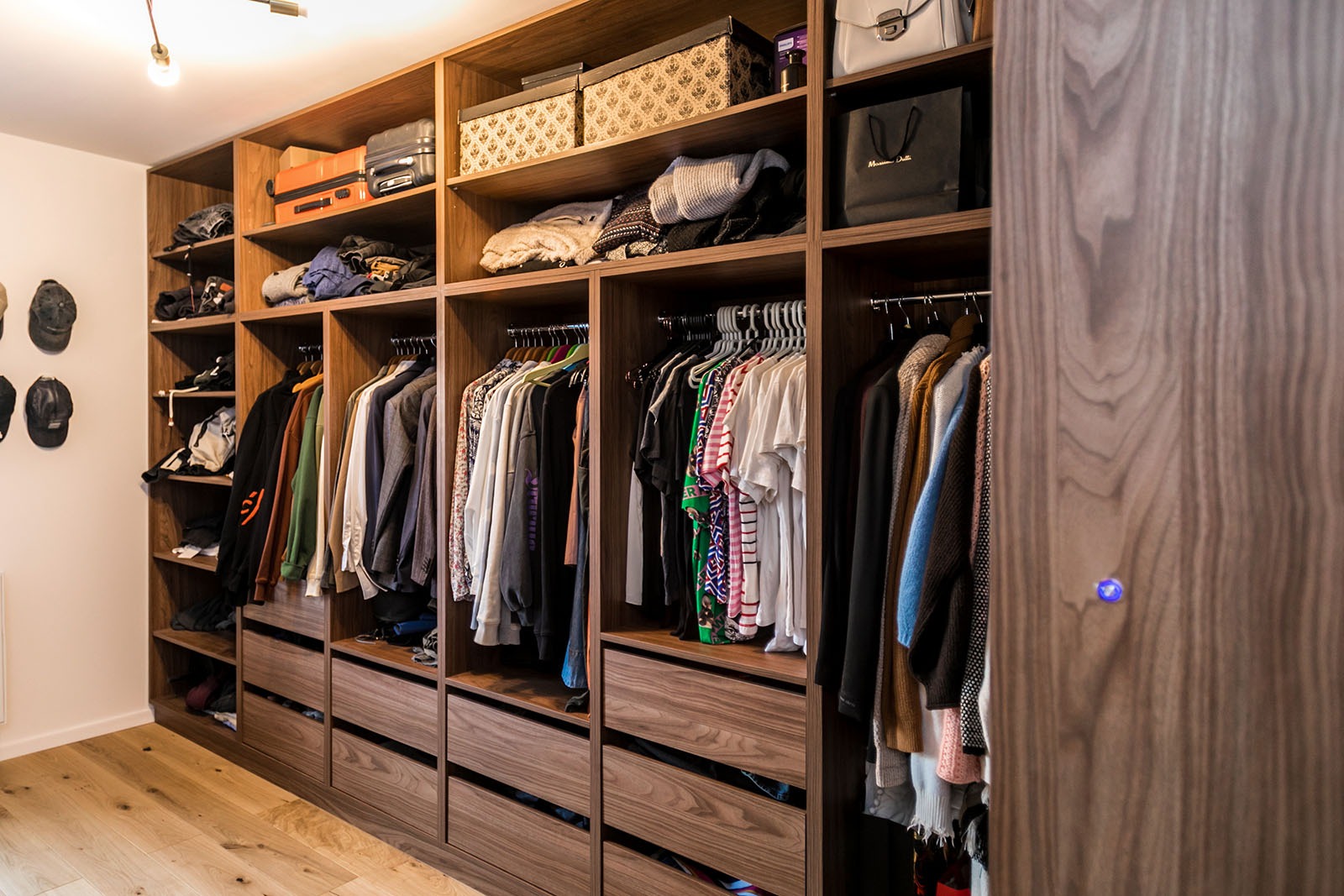 Pojemna szafa wnękowa z drewna orzechowego, z otwartymi półkami i szufladami, wypełniona ubraniami, torbami i pudełkami. Widoczny fragment drewnianej podłogi.