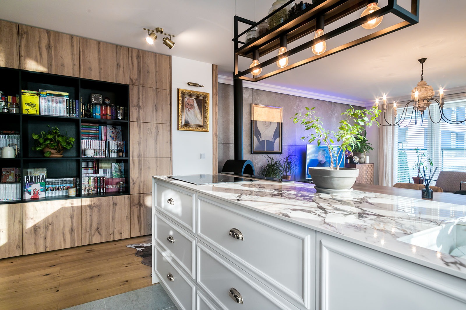 Nowoczesna kuchnia z wyspą i marmurowym blatem, drewniane szafki z czarnym regałem na książki, oświetlenie dekoracyjne i rośliny doniczkowe.