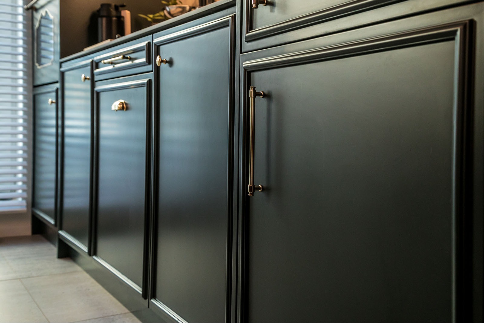 Ciemne, matowe fronty szafek kuchennych z frezowanymi obramowaniami i złotymi uchwytami, widoczne z bliska, z delikatnym odbiciem światła w tle.