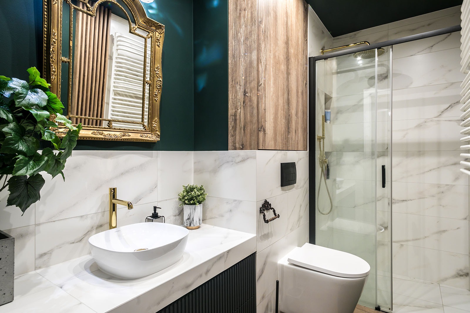 Elegancka łazienka z marmurowymi płytkami, złotą armaturą i drewnianą szafką. Umywalka nablatowa, lustro w ozdobnej ramie i kabina prysznicowa tworzą spójną całość.