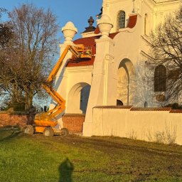 Kamil Rekowski - Prace na dachu kościoła z użyciem podnośnika koszowego. Widoczna biała fasada budynku i czerwona dachówka. W tle drzewa i błękitne niebo.